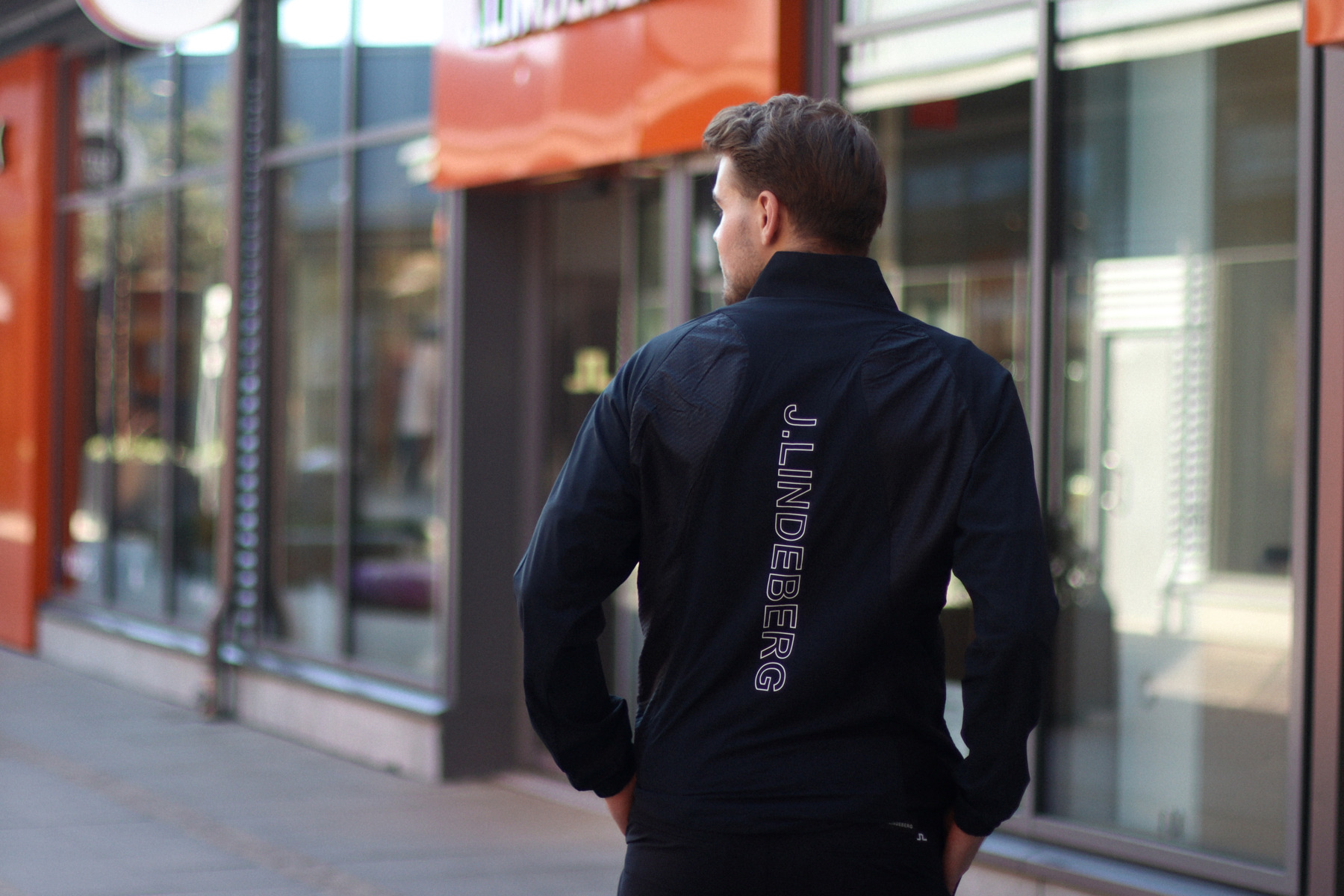 Helsinki Outlet village A person walks on a sidewalk beside a row of shops, wearing a black jacket with the text "J.LINDEBERG" printed vertically on the back.