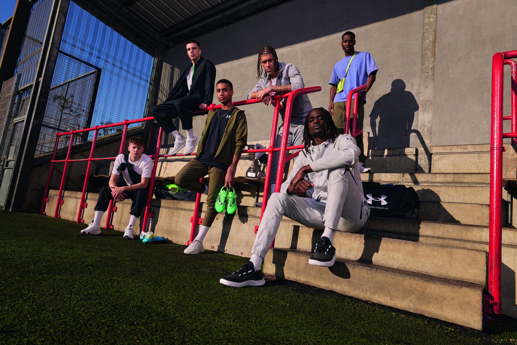 Kuusi nuorta miestä urheilullisissa katumuotiasuissa poseeraa punaisilla metallikaiteilla ja betoniportailla ulkoilmaurheilukentän laidalla. Aurinko paistaa matalalta luoden pitkiä varjoja. Yhdellä henkilöistä on kirkkaanvihreät jalkapallokengät kädessään, ja etualalla näkyy Under Armour -urheilukassi.